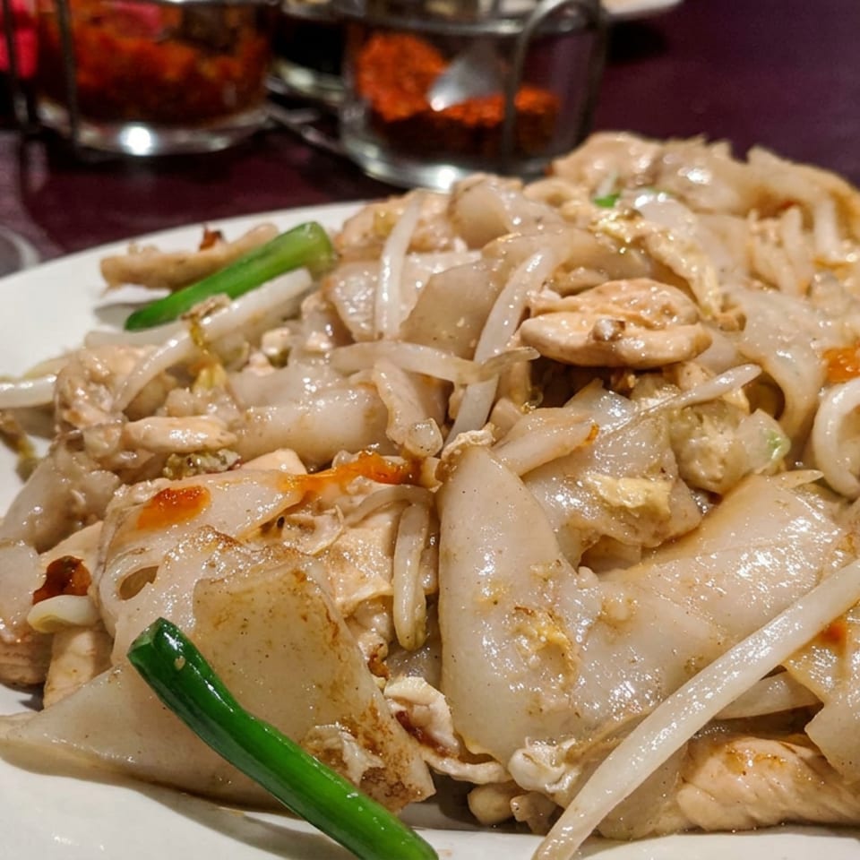 Plate of stir-fried noodles with chicken, bean sprouts, onions, green onions, and chili peppers on a restaurant table.