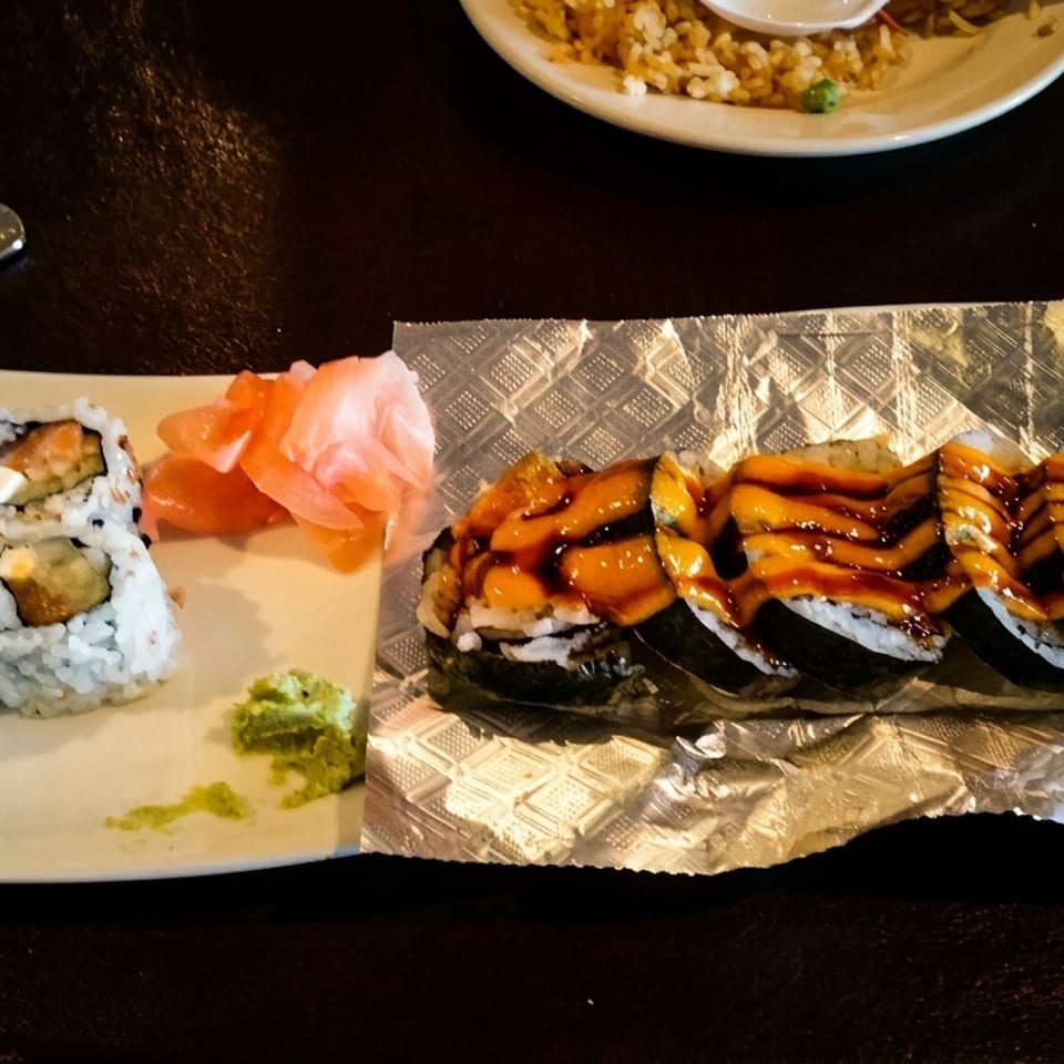 Sushi platter featuring assorted rolls, a glazed eel-topped roll, pickled ginger, wasabi, and fried rice on a dark table.
